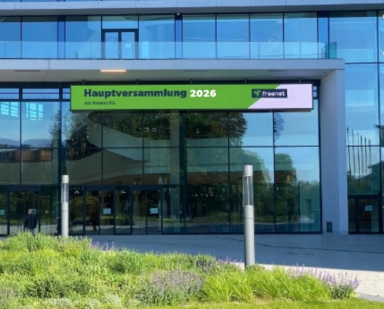 Eingang eines modernen Gebäudes mit großen Glasfenstern, darüber ein grünes Schild mit der Aufschrift ‚Hauptversammlung 2026 der Freenet AG‘.