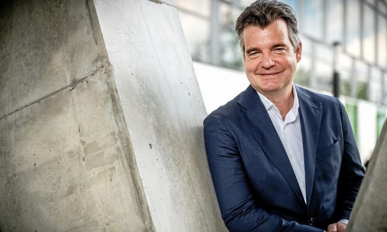 Mann in dunkelblauem Anzug lehnt sich lächelnd an eine große Betonstruktur in modernem Gebäude.