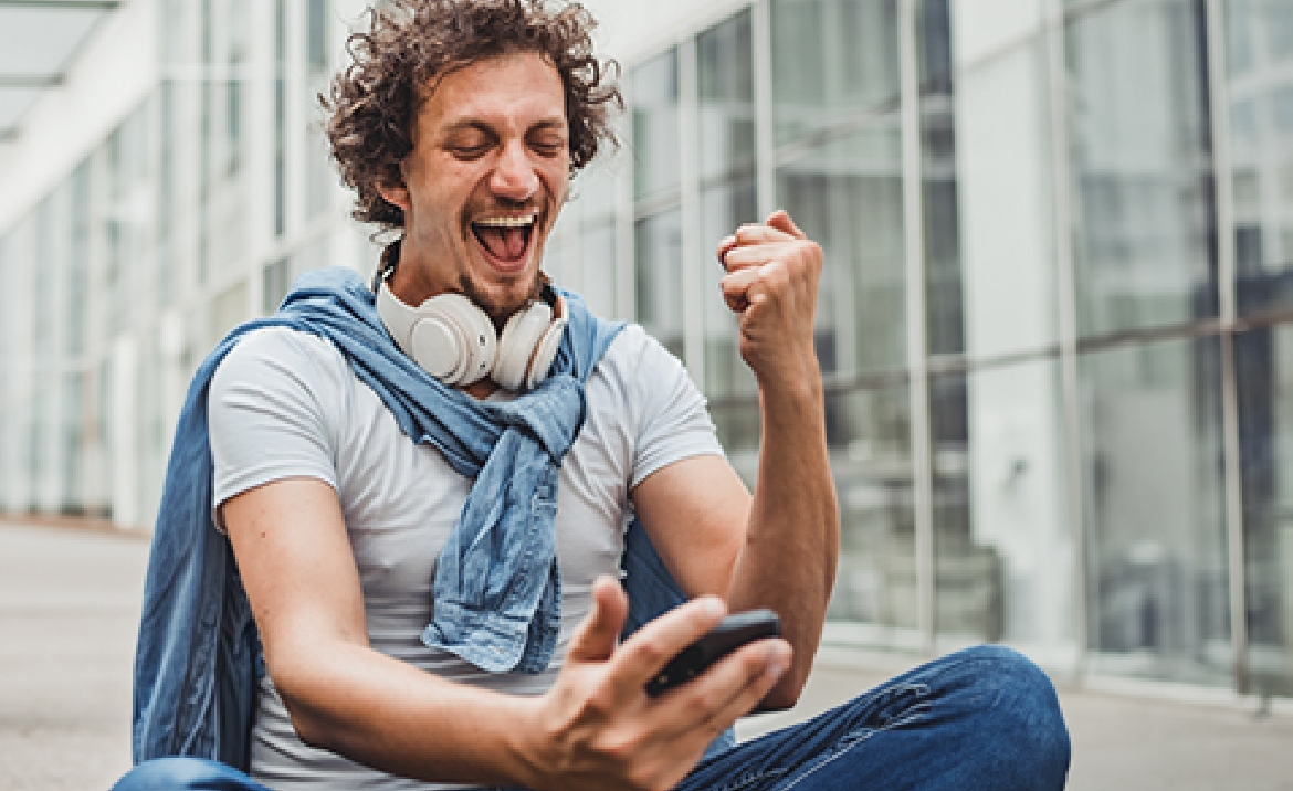 Junger Mann mit Kopfhörern zeigt freudige Geste und schaut auf Handy vor einem Gebäude mit Glaswand.