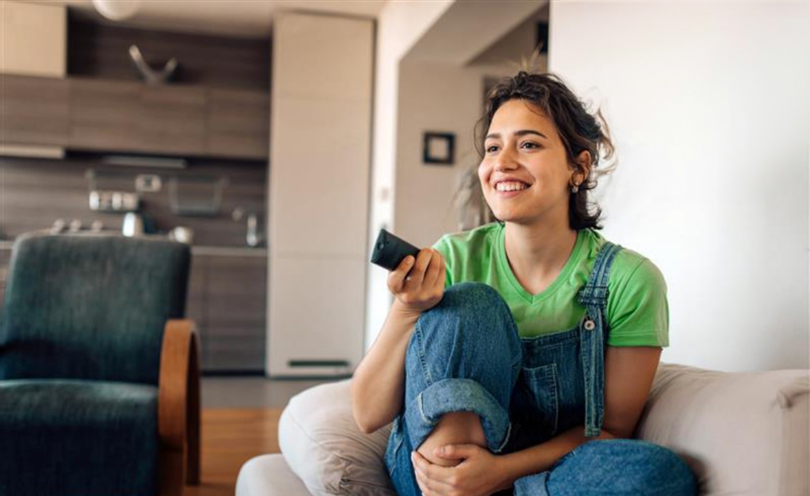 Frau in grüßem T-Shirt und Jeans sitzt lächelnd auf einem Sofa und hält eine Fernbedienung in einem modernen Wohnzimmer.